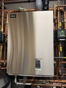 A stainless steel IBC VX high-efficiency wall mounted boiler mounted, surrounded  by copper piping, brass valves, and a Taco circulator pump for a heating system installed by a YYC ELECTRICIAN in Bragg Creek.